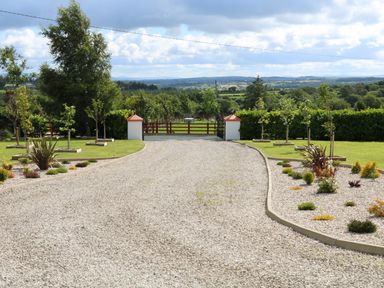 A gravel driveway with low garden beds and young trees on either side leading to red gates with countryside in the background at Sallywood House Carn near Ballybofey County Donegal