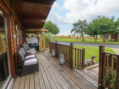 A wooden deck with four wicker chairs with cushions and a hot tub outside a cabin with a lawn and trees at Cornfield in Northallerton