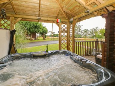 A jacuzzi with two wine glasses and a bottle under a wooden gazebo overlooking a grassy area at Cornfield in Northallerton