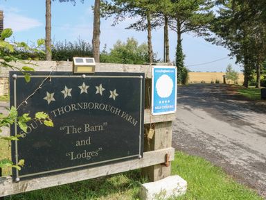 A black sign with gold text for south thornborough farm the barn and lodges beside a driveway with trees and fields at Cornfield in Northallerton