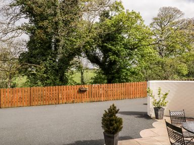 A paved patio area with two potted plants and metal chairs next to a wooden fence and trees at The Old Bothy in Bolton Low Houses near Wigton