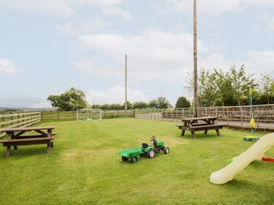 A fenced grass field with picnic tables a slide a toy tractor and a small soccer goal at The Old Grainstore in Bolton Low Houses near Wigton