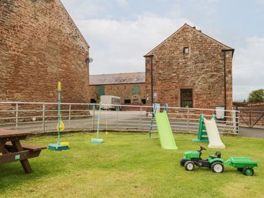A grass yard with two slides a toy tractor and outdoor games near stone buildings at The Old Grainstore in Bolton Low Houses near Wigton