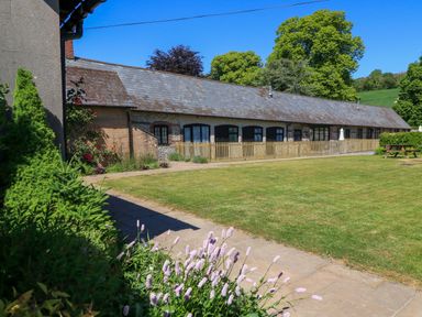 A long brick and flint building with multiple doors and windows a grass lawn and flowers at The Old Stables in Winterborne Stickland
