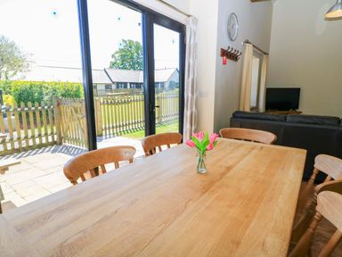 A dining area with a wooden table and chairs a vase with flowers on the table large glass doors overlooking a garden and fence a black sofa and television set at The Old Stables in Winterborne Stickland