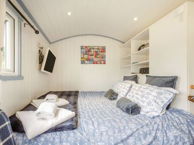 A small bedroom with a blue patterned bedspread white pillows shelves a wall-mounted TV and folded towels at The Shepherd's Hut in Aberdovey