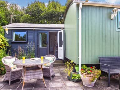 An outdoor patio with a wicker table and chairs a black bench and flower pots outside two small buildings at The Shepherd's Hut in Aberdovey