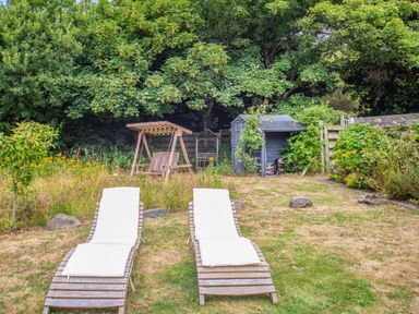 A garden with two wooden lounge chairs with cushions a wooden swing and a small shed surrounded by trees and plants at The Shepherd's Hut in Aberdovey