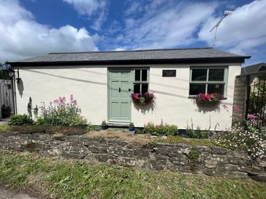 A small building with a green door and two windows with flower boxes a stone wall and garden in front at Palmers Lodge in Egloskerry near Launceston