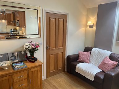 A small living room with a wooden door a brown leather sofa with pillows a wooden cabinet with flowers and a mirror above at Palmers Lodge in Egloskerry near Launceston