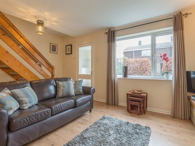 A living room with a brown leather sofa and pillows a gray rug wooden stairs and a window with curtains at Crumstone Corner in Seahouses