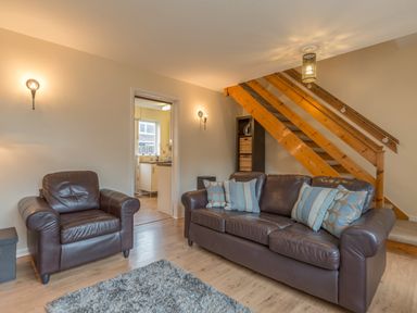 A living room with a brown leather sofa and armchair near wooden stairs and an open door to a kitchen at Crumstone Corner in Seahouses
