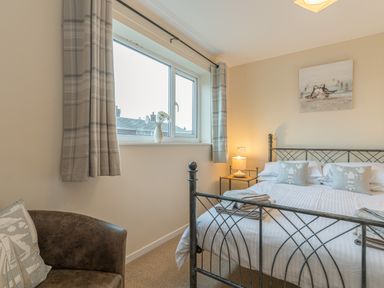 A bedroom with a bed metal frame a window with curtains a chair and a bedside table with a lamp at Crumstone Corner in Seahouses