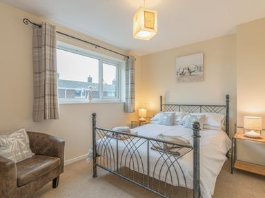 A bedroom with a metal bed frame a brown armchair bedside tables with lamps and a window with curtains at Crumstone Corner in Seahouses