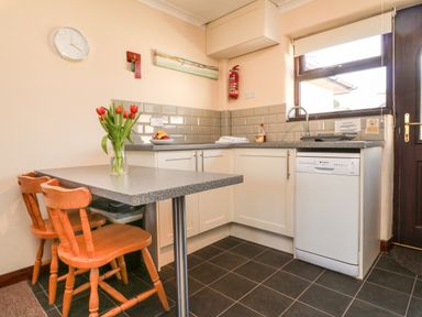 A kitchen with a table and two wooden chairs a vase with red flowers on the table a countertop with a sink dishwasher and a window at Honeysuckle Cottage in Bude