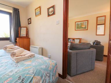 A bedroom with a bed covered in patterned bedding and folded towels next to a window and a dresser with a mirror and an adjoining room with two grey armchairs and framed pictures at Honeysuckle Cottage in Bude