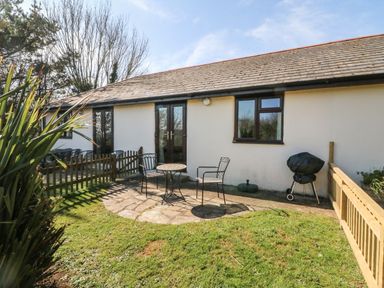 An outdoor patio area with a small round table and two chairs next to a white building with a window and double doors at Honeysuckle Cottage in Bude