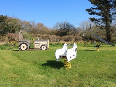 An outdoor playground with a wooden tractor, two spring riders shaped like sheep, and a metal slide at Honeysuckle Cottage in Bude