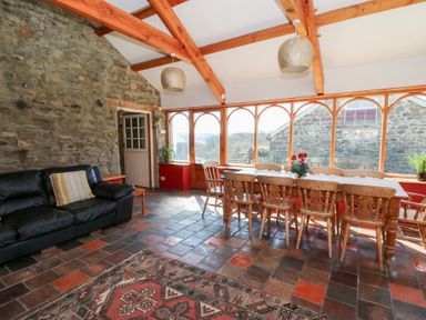 A dining area with wooden chairs and table next to large windows and a black sofa in a room with exposed stone wall and wooden beams at The Farmhouse in Aberaeron