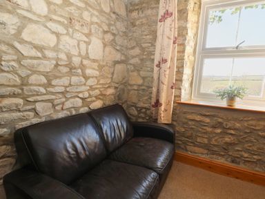 A corner with a black leather sofa next to a window with floral curtains and a potted plant on the windowsill at The Farmhouse in Aberaeron