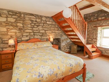A bedroom with a wooden bed and floral bedding under wooden stairs with stone walls at The Farmhouse in Aberaeron