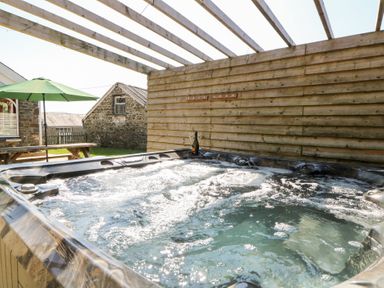 An outdoor hot tub with bubbling water under a wooden pergola and a picnic table with a green umbrella near stone buildings at The Farmhouse in Aberaeron