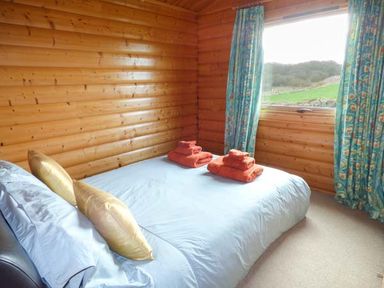 A bedroom with a double bed covered in light blue bedding and orange towels on the bed beside a window with blue patterned curtains in a wooden cabin at Ty Pren in Llanrhian near St Davids