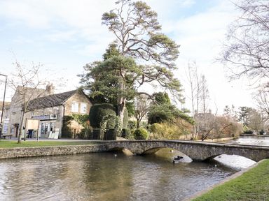 A stone arch bridge over a river with a stone building and trees in the background at Greyhound Barn in Barnsley near Cirencester