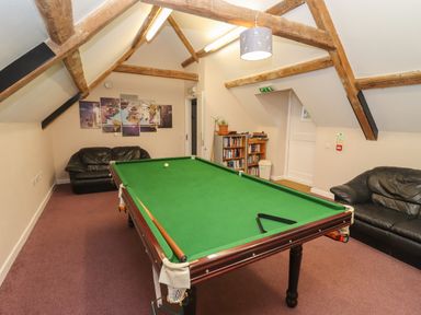 A game room with a pool table two black leather sofas bookshelves and wooden beams on the ceiling at Greyhound Barn Barnsley near Cirencester