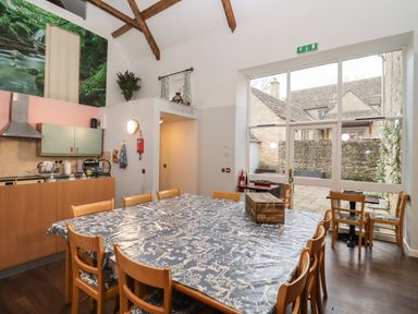 A dining area with a large table covered in a patterned tablecloth and wooden chairs near a kitchen and large window at Greyhound Barn Barnsley near Cirencester