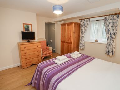 A bedroom with a bed covered with a purple striped blanket towels on top a wooden wardrobe chest of drawers with a TV and an armchair near a window with floral curtains at Greyhound Barn Barnsley near Cirencester