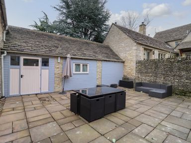 An outdoor patio area with stone paving a black glass top table with black wicker chairs a black wicker sofa a folded umbrella and stone buildings at Greyhound Barn Barnsley near Cirencester