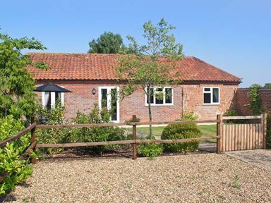 A single-story brick house with a tiled roof a wooden fence a gravel driveway and garden with trees and shrubs at Moat Farm Cottage in Wood Dalling