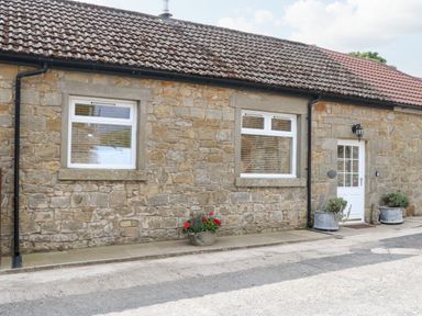 Stone cottage exterior with two windows and a white door with flower pots at Stable Cottage in Belford near Bamburgh