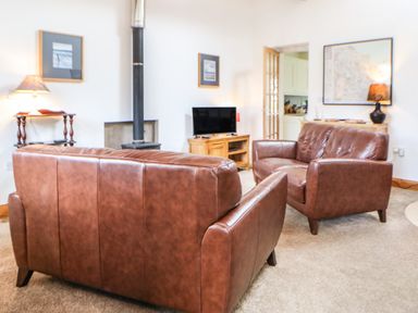 A living room with two brown leather sofas a TV on a wooden stand a wood burning stove and lamps at Stable Cottage Belford near Bamburgh