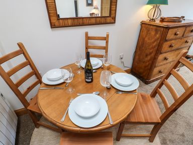 A wooden dining table set with white plates bowls utensils and wine glasses with four wooden chairs around and a wooden dresser with a lamp in a room at Stable Cottage in Belford near Bamburgh
