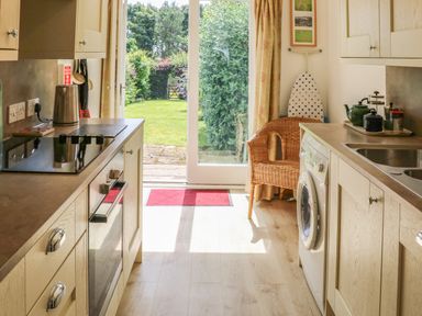 A kitchen with light wood cabinets a washing machine a wicker chair and a glass door leading to a garden at Stable Cottage Belford near Bamburgh