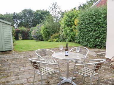 An outdoor patio with a round table and four chairs with a bottle and two glasses on the table and garden with shed and shrubs at Stable Cottage in Belford near Bamburgh