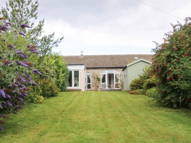 A garden with lawn, shrubs, flowers, a shed, and a house with patio doors at Stable Cottage in Belford near Bamburgh