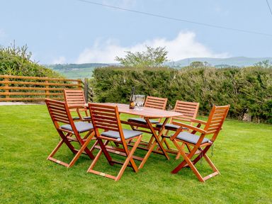 A wooden outdoor dining table with six chairs on grass with hedges and hills in the background at Pen Y Gaer in Llannefydd near Abergele and Denbigh