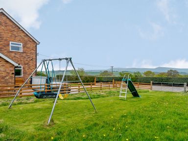 A backyard with a swing set and a slide next to a brick house with a wooden fence and green fields at Pen Y Gaer in Llannefydd near Abergele and Denbigh