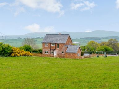 A brick house with a fenced yard and swing set surrounded by green fields and hills in the background at Pen Y Gaer in Llannefydd near Abergele and Denbigh