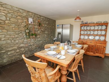 A dining room with a wooden table set for a meal wooden chairs stone wall with hanging plants wooden cabinet with plates and a refrigerator at Mill Cottage in Aberaeron