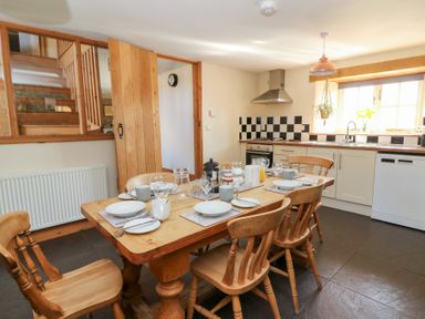 A dining area with a wooden table set for six and a kitchen with black and white tiled backsplash at Mill Cottage in Aberaeron