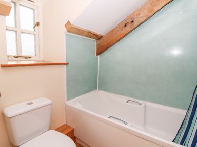 A bathroom with a white toilet under a window and a bathtub with handles and a sloped ceiling with a wooden beam at Mill Cottage in Aberaeron
