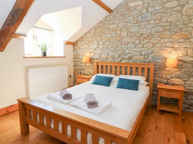A bedroom with a wooden bed frame white bedding stone wall two bedside tables with lamps and a window with a plant at Mill Cottage in Aberaeron