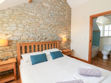 A bedroom with a wooden bed frame against a stone wall with bedside tables and lamps and an open door showing a bathroom at Mill Cottage in Aberaeron
