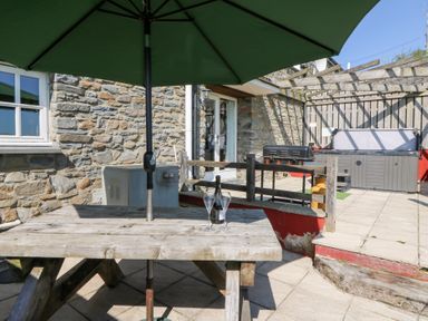 An outdoor patio with a wooden picnic table and green umbrella a bottle and two glasses on the table a stone wall a barbecue and a hot tub under a wooden pergola at Mill Cottage in Aberaeron