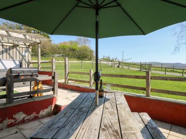 An outdoor patio with a wooden picnic table and umbrella a bottle and two glasses on the table and a barbecue grill with a fence and grassy field beyond at Mill Cottage Aberaeron
