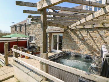 An outdoor patio with a hot tub under a wooden pergola and a BBQ grill near a stone house at Mill Cottage in Aberaeron